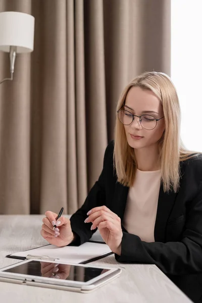 Aantrekkelijke blonde vrouw in glazen en een Business Suit werken op een Tablet, praten over de telefoon en het maken van aantekeningen in een notebook. Hotel kamer, tafel bij het raam. — Stockfoto