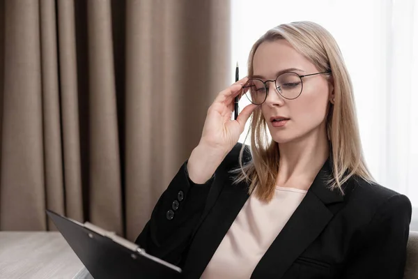 Attraktive blonde Frau mit Brille und Businessanzug, die an einem Tablet arbeitet, telefoniert und Notizen in einem Notizbuch macht. Hotelzimmer, Tisch am Fenster. — Stockfoto