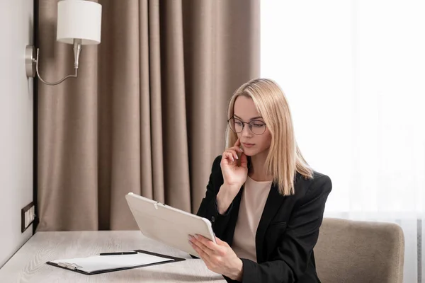 Atraktivní blondýnka v brýlích a firemní oblek pracující na tabletu, mluví na telefonu a berou si poznámky v poznámkovém bloku. Hotelový pokoj, stůl u okna. — Stock fotografie