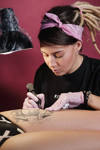 Female tattoo master tattooing female client. Figure nuns on the thigh. Black tattoo machine and lamp. Workplace artist.