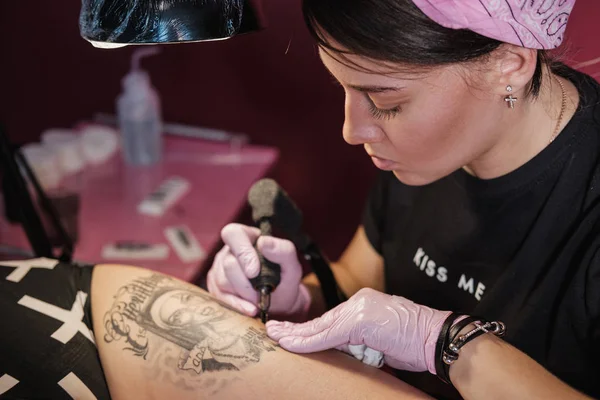 Female tattoo master tattooing female client. Figure nuns on the thigh. Black tattoo machine and lamp. Workplace artist.