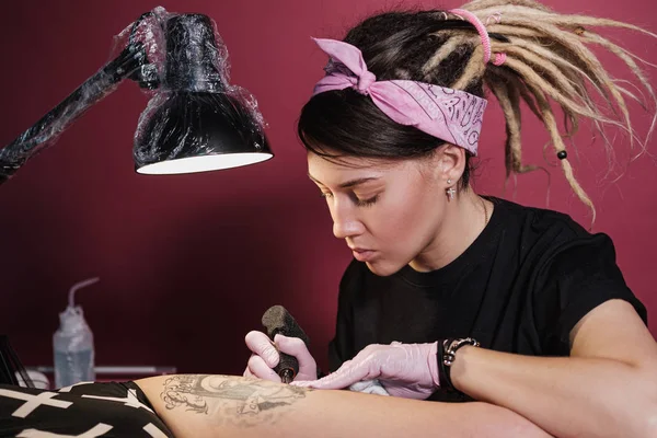 Tatuagem feminina mestre tatuando cliente feminino. Imagina freiras na coxa. Máquina de tatuagem preta e lâmpada. Artista local de trabalho . — Fotografia de Stock