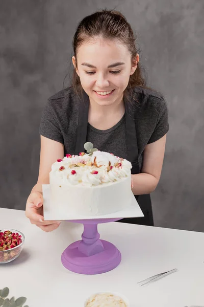 Atractiva joven morena pastelera presenta un pastel blanco con pequeñas flores rojas de una rosa de comida . —  Fotos de Stock