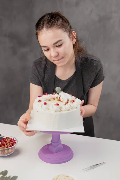 Attractive young woman brunette confectioner presents a white cake with small red flowers of a food rose. — Stock Photo, Image