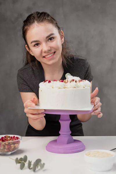 Jolie jeune femme brune confiseur présente un gâteau blanc avec de petites fleurs rouges d'une rose alimentaire . — Photo