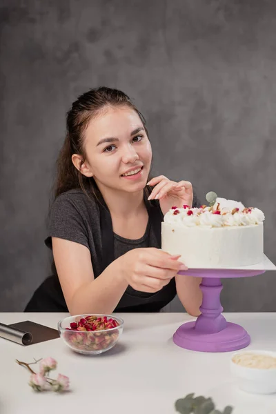 Attractive young woman brunette confectioner presents a white cake with small red flowers of a food rose.