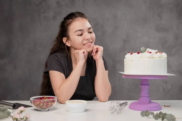 Atractiva joven morena pastelera presenta un pastel blanco con pequeñas flores rojas de una rosa de comida . —  Fotos de Stock