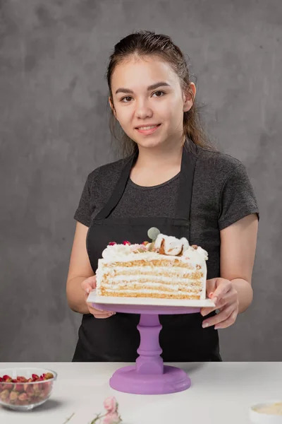 La giovane donna attraente il pasticciere bruna presenta una torta bianca con piccoli fiori rossi di una rosa di cibo . — Foto Stock