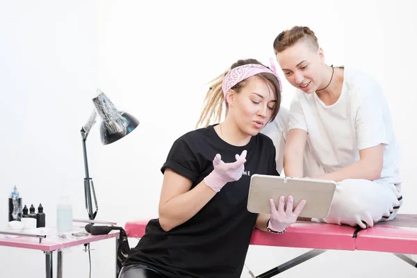 Tatuagem profissional mulher mestre mostra esboços no tablet para a menina cliente. Discussão futuras tatuagens . — Fotografia de Stock