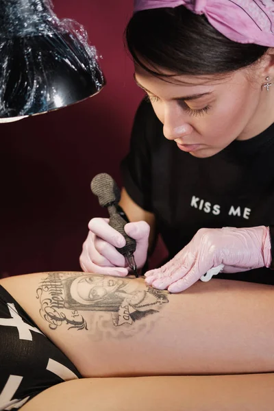 Female tattoo master tattooing female client. Figure nuns on the thigh. Black tattoo machine and lamp. Workplace artist.