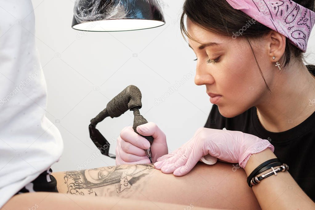 Female tattoo master tattooing female client. Figure nuns on the thigh. Black tattoo machine and lamp. Workplace artist.