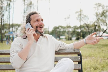 Kuyruk uzun saçlı yaşlı bir yakışıklı adam duygusal bir bankta bir parkta bir cep telefonu konuşuyor.