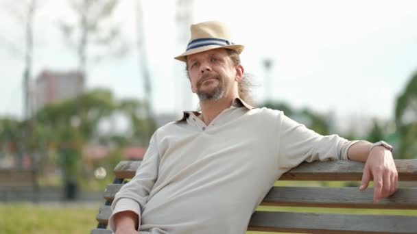 帽子に長い白髪のリラックスした魅力的な中年の男性が座って、公園のベンチに座って休みます。夏の夜、暑さ. — ストック動画