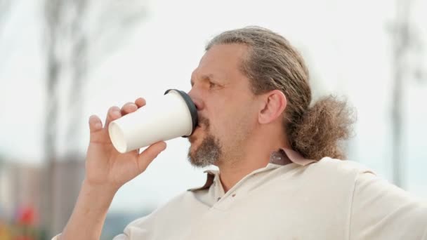 Um homem de meia-idade relaxado e atraente com cabelos longos e grisalhos bebe café de um copo descartável em um banco em um parque verde . — Vídeo de Stock