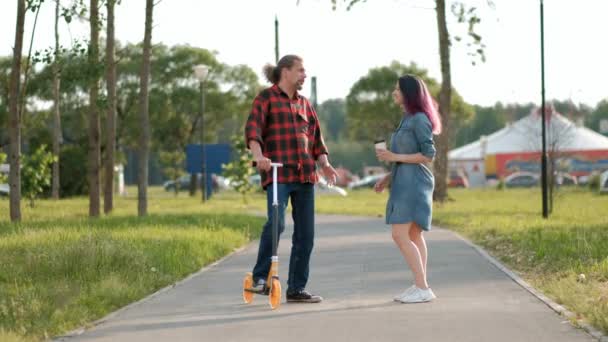 Een ontspannen aantrekkelijke man van middelbare leeftijd met lang grijs haar op een kick-scooter en een jonge vrouw met geverfd haar begroeten en omarmen. — Stockvideo