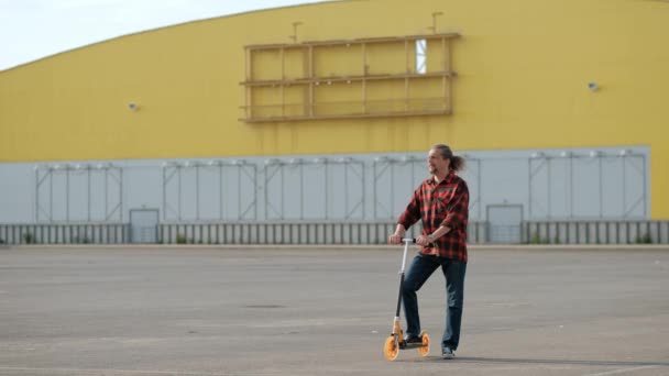 Rilassato attraente uomo di mezza età con lunghi capelli grigi in una camicia rossa a scacchi cavalca su un kick-scooter . — Video Stock