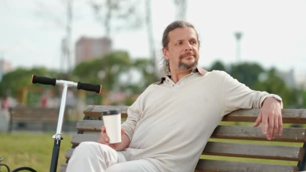 Un uomo rilassato e attraente di mezza età con lunghi capelli grigi beve caffè da un bicchiere usa e getta su una panchina in un parco verde . — Video Stock