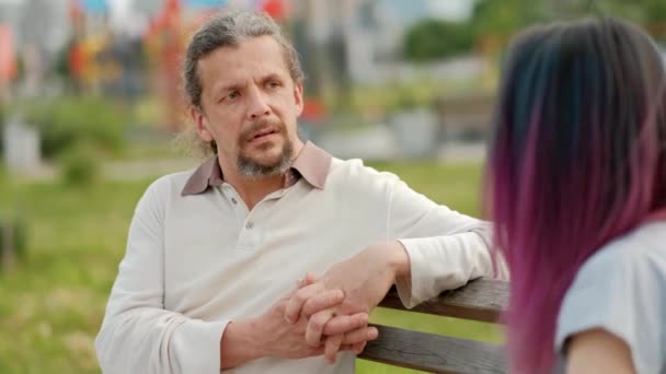 Um homem de meia-idade atraente descontraído com cabelos grisalhos longos e uma jovem mulher com cabelo tingido sentada em um banco de parque e conversando . — Vídeo de Stock