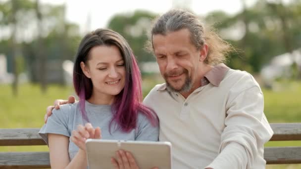 Relajado atractivo hombre de mediana edad con el pelo largo y gris y una mujer joven con el pelo teñido abrazando y viendo fotos en una tableta . — Vídeos de Stock