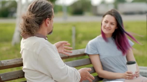 Poklidně přitažlivý muž středního věku s dlouhými šedými vlasy a mladou ženou s barvýma vlasama, sedící na lavičce v parku a žvaničení. — Stock video