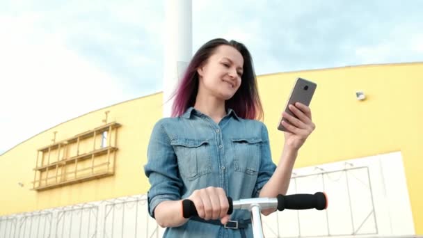 Jolie jeune femme avec les cheveux teints dans une robe en denim et des baskets blanches chevauchant un kick-scooter et en utilisant un smartphone . — Video
