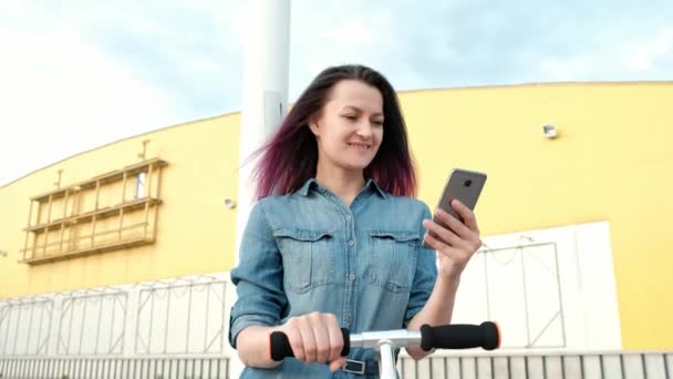 Attraktive junge Frau mit gefärbten Haaren im Jeanskleid und weißen Turnschuhen auf einem Tretroller und mit einem Smartphone. — Stockvideo