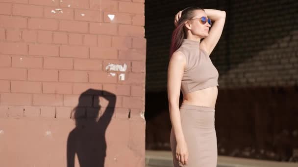 Νεαρή όμορφη γυναίκα σε μια φούστα και γυαλιά ηλίου σε ένα φόντο τοίχου τούβλο. Ο άνεμος χτενίζει τα μαλλιά. — Αρχείο Βίντεο