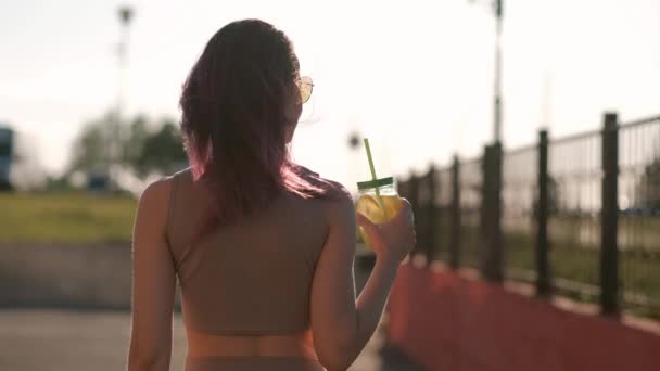 Jonge mooie vrouw in zonnebrillen is het drinken van een cocktail uit een glazen pot met een stro op een straat in de stad. — Stockvideo