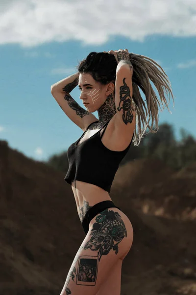 Krásná sexy žena v černém spodním prádle. Tělo je pokryté mnoha tetování. Strašné zámky na hlavě. Póziva proti písečné kariéře a černé tkanině. — Stock fotografie