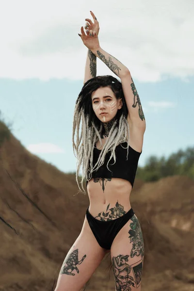 Hermosa mujer sexy en ropa interior negra. El cuerpo está cubierto de muchos tatuajes. Ricitos en la cabeza. Posando contra la carrera de arena y tela negra . — Foto de Stock