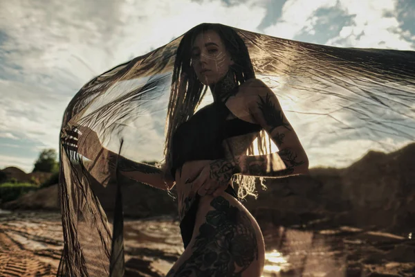 Siyah iç çamaşırı güzel seksi kadın. Vücut birçok dövmeler ile kaplıdır. Kafasından dreadlocks. Kumlu kariyer ve siyah kumaş karşı poz.