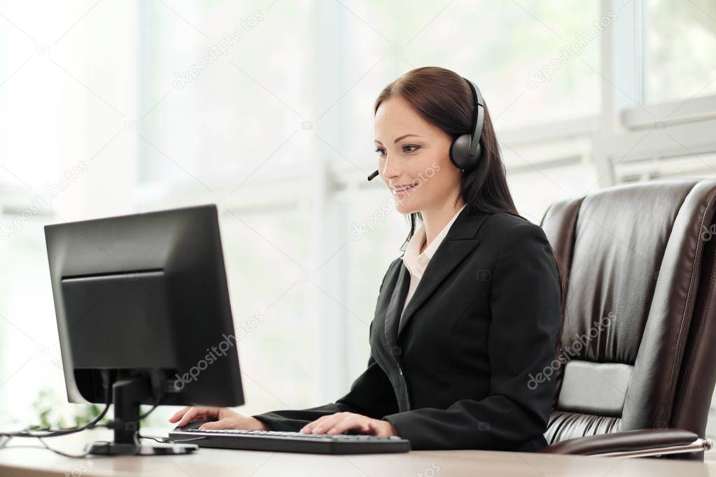 A young attractive woman wearing a black jacket in a black jacket is sitting at her desk in a executive chair. Holds a video conference in headphones with a microphone.