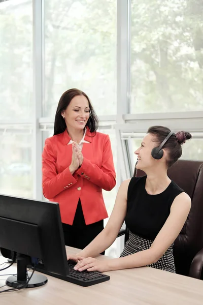 Two young women work in a bright office at the computer. Discuss workflows and enjoy a successful deal. Head and subordinate.