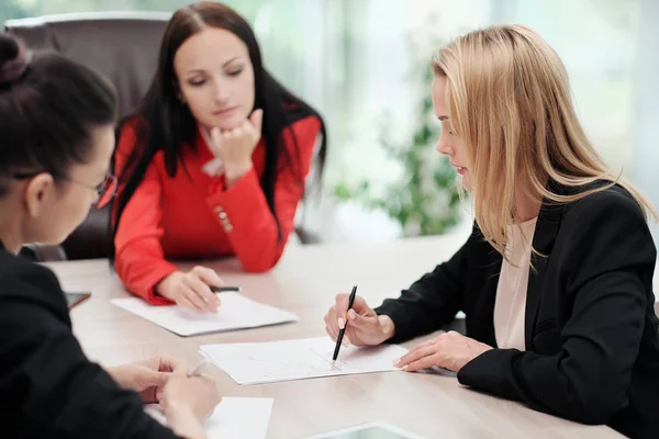 Tres mujeres jóvenes y atractivas en trajes de trabajo están sentadas en un escritorio y discutiendo los flujos de trabajo. Cabeza y subordinados. Equipo de trabajo de profesionales y colegas . —  Fotos de Stock