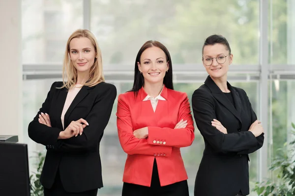 Drie jonge aantrekkelijke vrouwen in business suits poseren tegen de achtergrond van een licht kantoor. Hoofd en ondergeschivelde. Werk team van professionals en collega's. — Stockfoto