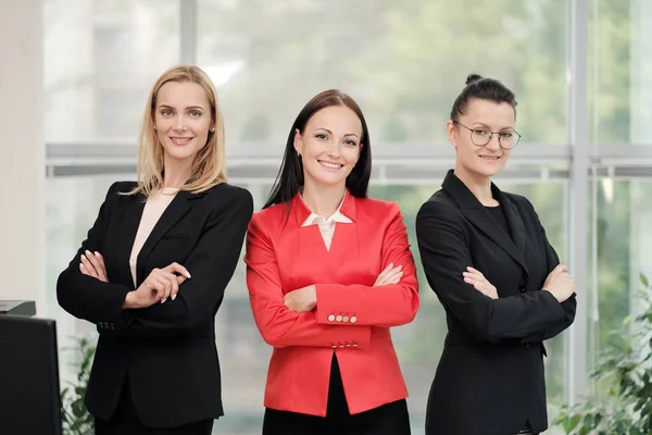 Drie jonge aantrekkelijke vrouwen in business suits poseren tegen de achtergrond van een licht kantoor. Hoofd en ondergeschivelde. Werk team van professionals en collega's. — Stockfoto