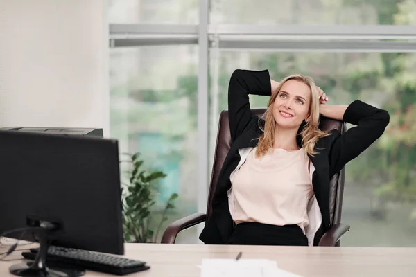 Een jonge aantrekkelijke vrouw in Business Suit zit aan een bureau. Strekt zich uit en ontspant op stoel. Voeten zijn op tafel. Rest van het werk. Vermoeidheid, onderbreking in het werk. — Stockfoto