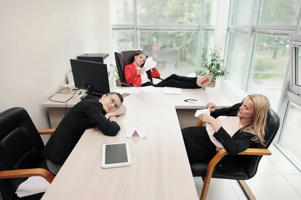 Tres hermosas chicas jóvenes en trajes de negocios están sentadas en el escritorio de la oficina. Cansado del trabajo. Tira aviones de papel y ríete. Descanso y descanso . —  Fotos de Stock