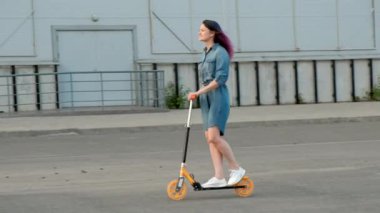 Bir Denim elbise ve şehir sokaklarında bir Kick Scooter sürme beyaz spor ayakkabıları boyalı saçlı çekici genç kadın.