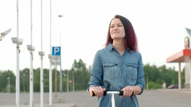 Attraktiv ung kvinna med färgat hår i en Jeansklänning och vita sneakers som rider på en spark skoter genom gatorna i staden. — Stockvideo