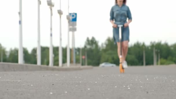 Attraktiv ung kvinna med färgat hår i en Jeansklänning och vita sneakers som rider på en spark skoter genom gatorna i staden. — Stockvideo