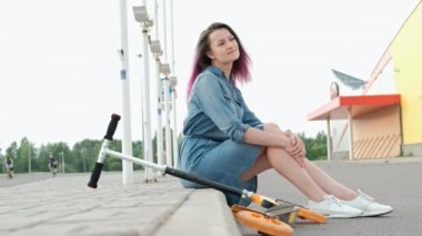 Kot elbise ve beyaz spor ayakkabıları boyalı saçlı çekici genç kadın kaldırımda oturur ve dayanır.