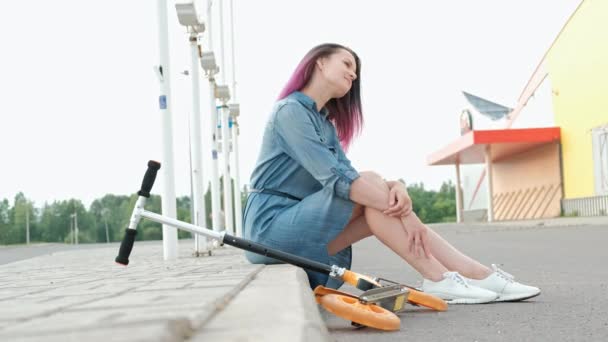 Attraktiv ung kvinna med färgat hår i en Jeansklänning och vita sneakers sitter på trottoarkanten och vilar. — Stockvideo