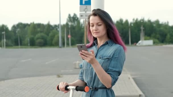 Jolie jeune femme avec les cheveux teints dans une robe en denim et des baskets blanches chevauchant un kick-scooter et en utilisant un smartphone . — Video