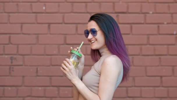 Güneş gözlüğü genç güzel kadın bir tuğla duvar ile bir evin arka planına karşı bir saman ile cam bir kavanozdan limonata kokteyl içiyor. — Stok video