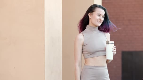 Une jeune belle femme aux cheveux teints de vert et violet dans des lunettes de soleil boit une boisson chaude d'une tasse thermique blanche sur le fond d'une maison avec un mur de briques . — Video