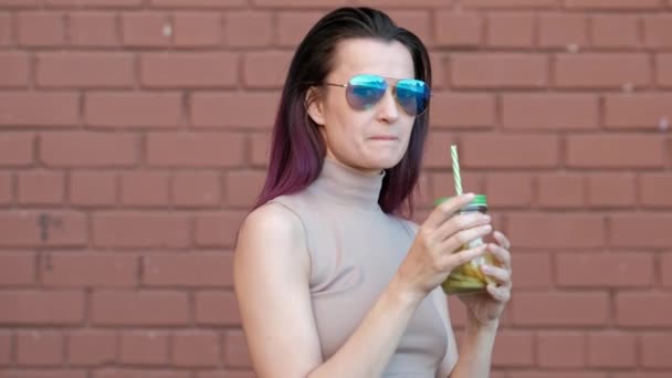 Joven hermosa mujer en gafas de sol está bebiendo cóctel de limonada de un frasco de vidrio con una paja sobre el fondo de una casa con una pared de ladrillo . — Vídeo de stock