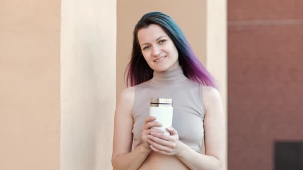 一个戴着太阳镜，头发染成绿色和紫色的年轻漂亮女人正在一座有砖墙的房子背景下喝着白色热水瓶。. — 图库视频影像