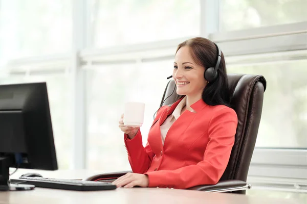 Mladá atraktivní žena v červeném saku sedí na stole u vedoucího výkonné kanceláře v červeném plášti. Pořádá videokonferenci ve sluchátkách s mikrofonem. Popíjení kávy z bílého poháru. — Stock fotografie