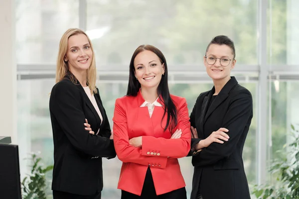 Drie jonge aantrekkelijke vrouwen in business suits poseren tegen de achtergrond van een licht kantoor. Hoofd en ondergeschivelde. Werk team van professionals en collega's. — Stockfoto